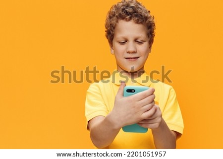 a pensive, cute boy with curly hair stands holding his smartphone in his hand on a yellow background with an empty space for an advertising mockup