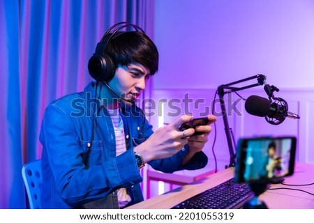 Playing video games on smartphone. Young asian handsome man sitting on chair holding cellphone in his hand. Exited streamer wearing headset in online mobile game in neon room .Esport streaming game.