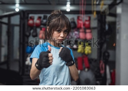 A determined athletic woman takes a picture with an intense look on her face while holding a thumbs up. Boxing gloves background.