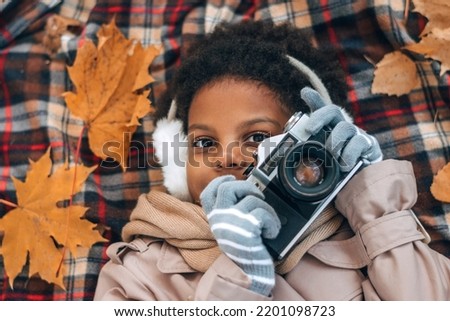 Cute African-American girl in fur headphones takes pictures with a camera lying on a blanket in an autumn park.Diversity,autumn concept.