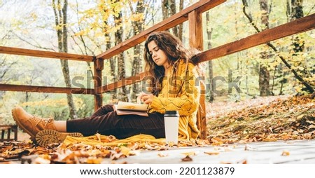  Brunette woman in yellow sweater sitting on a fallen autumn leaves in a park, reading a book or write a diary