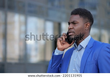 African American man has call with colleague on black smartphone. Bearded businessman talks on phone with worker against office building