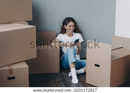 Happy latina female uses mobile phone apps, choosing moving service, preparing for relocation, answer her mover online, sitting on the floor with packed things in cardboard boxes. Easy moving day Royalty-Free Stock Photo #2201172827