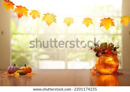Autumn home decoration. Pumpkin lantern for Thanksgiving table setting. Orange leaves and flowers arrangement in living room. Fall colors in house decor.  Decorated wooden table at big window.