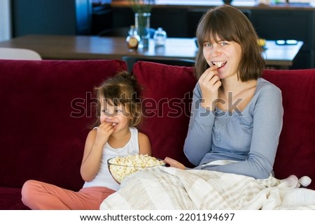 Mom and daughter are sitting on a bright sofa and eating popcorn together. They are covered with a warm blanket. The concept of comfort, love and mutual understanding in the family