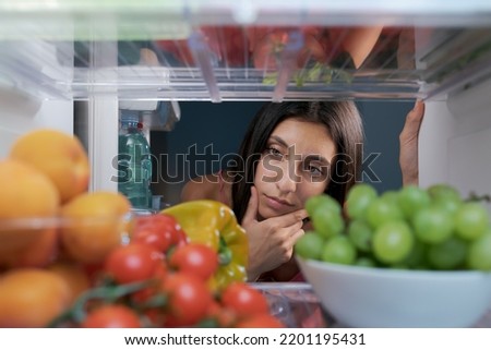 Woman looking in the fridge and thinking with hand on chin, fresh fruit and vegetables in the foreground, POV shot from inside of the fridge Royalty-Free Stock Photo #2201195431