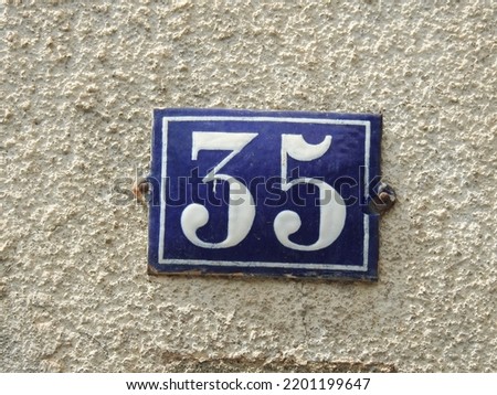 House number (address) thirty five (35) sign, white numbered blue plate (closeup) against white wall of old building background. Grunge, texture, wallpaper