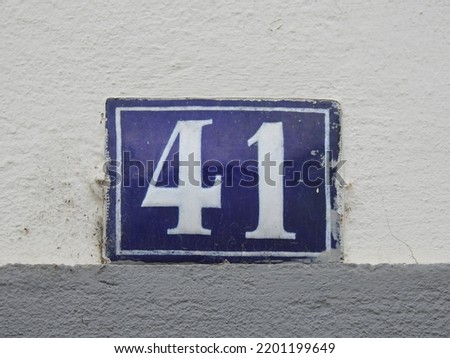 House number (address) forty one (41) sign, white numbered blue plate (closeup) against white wall of old building background. Grunge, texture, wallpaper