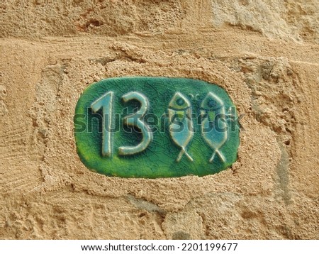 House number (address) thirteen (13) sign, green-turquoise numbered ceramic plate (closeup) with Pisces zodiac symbol against brick wall of old building background in Jaffa Israel. Grunge, texture