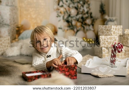 Cute blond toddler preschool boy, reading a book and opening present on Christmas on cozy home, lights and decoration