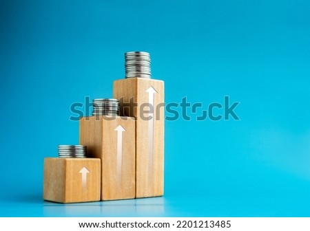 Shining rise up arrow on wooden cube blocks top with coins stacked, bar graph chart steps on blue background with space, investment, income, inflation, business growth, economic improvement concepts.
