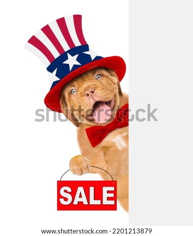 Happy Mastiff puppy wearing like Uncle Sam looks from behinde empty white banner and shows signboard with labeled "sale". isolated on white background