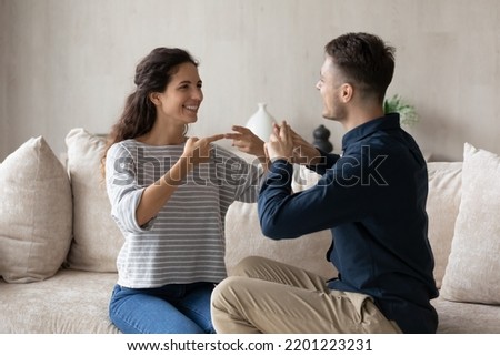 Smiling young woman who is deaf and man using sign language, sitting on couch at home, excited beautiful girlfriend and boyfriend having fun, enjoying pleasant conversation, hard of hearing