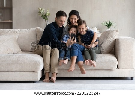 Happy parents and children having fun with smartphone at home, smiling mother and father with kids looking at smartphone screen, sitting on cozy couch, watching cartoons or making video call