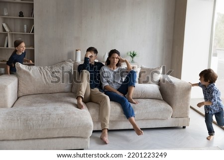 Tired parents sitting on couch at home while noisy little kids running and shouting around, young mother and father suffering from headache, feeling exhausted and annoyed by naughty active children Royalty-Free Stock Photo #2201223249