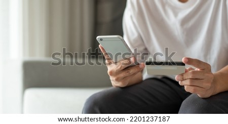Young man in a white shirt sits and using smartphone and hand holding credit card for pay online shopping on the couch at home. She smiled happily not having to leave the house to go shopping. Royalty-Free Stock Photo #2201237187