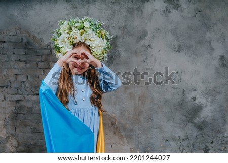 Little girl in flower wreath standing with Ukraininan flag against loft wall showing heart. Blank space for text.
