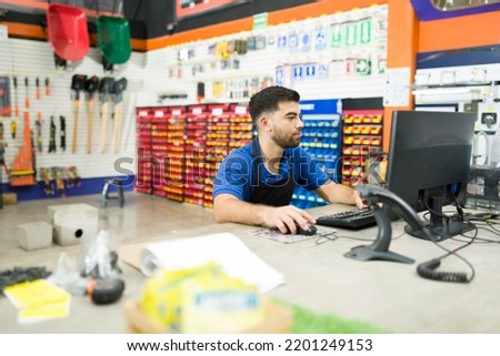 Busy young man employee working as a cashier at the hardware shop and using the computer to find a product or checking the inventory Royalty-Free Stock Photo #2201249153