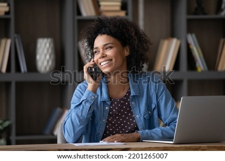 Happy young African American woman involved in mobile phone call conversation, distracted from computer work, discussing issues distantly, listening good news, sitting at table at home office. Royalty-Free Stock Photo #2201265007