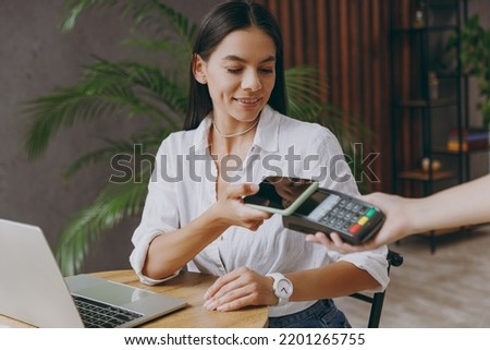 Young happy woman wears white shirt hold pay waiter hold mobile cell phone wireless bank payment terminal waiter sit at table in coffee shop cafe restaurant indoor work or study on laptop pc computer Royalty-Free Stock Photo #2201265755