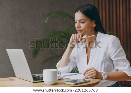 Young minded latin IT woman wear white shirt use work or study on laptop pc computer hold pen write sit alone at table in coffee shop cafe restaurant indoors. Freelance mobile office business concept Royalty-Free Stock Photo #2201265763