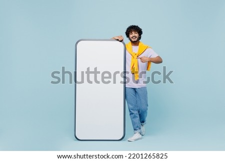 Full size young bearded Indian man 20s wears white t-shirt stand near pointing on big mobile cell phone with blank screen workspace area isolated on plain pastel light blue background studio portrait