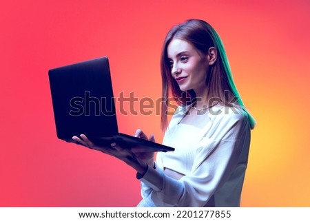 Portrait of young woman posing, having video conference on laptop isolated over red background in neon light. Online working. Concept of beauty, lifestyle, youth, emotions, facial expression, ad.