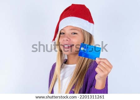 Close up photo of optimistic little kid girl with Christmas hat wearing yarn jacket over white background hold card