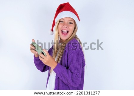 Nice addicted cheerful little kid girl with Christmas hat wearing yarn jacket over white background using gadget playing network game
