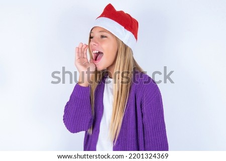 little kid girl with Christmas hat wearing yarn jacket over white background look empty space holding hand face and screaming or calling someone.