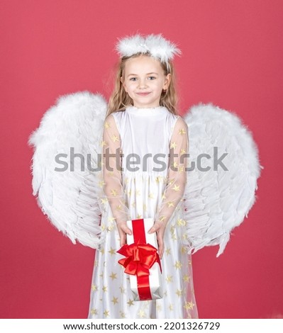 Child angel little girl with present gift, studio portrait. Little angel with white wings holds gift. Valentine gift card. Cute angel child girl with angels wings, isolated on red. Valentines day. Royalty-Free Stock Photo #2201326729