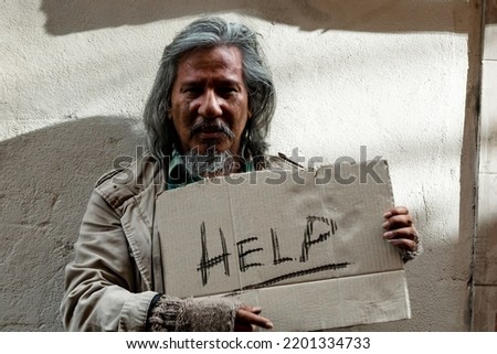 A long-haired Asian man, dirty dressed as a homemaker, holds up a paper placard that reads for help from a passerby because he has no food, money, and sleeps on the roadside.