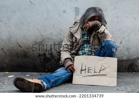 homeless, long-haired Asian man sits hopelessly leaning against a wall as there is no one to help him with work and food in his hand holding a sign for help. Homeless sleep on streets