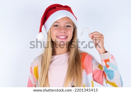 little kid girl with Christmas hat wearing yarn jacket over white background holding an invisible braces aligner, recommending this new treatment. Dental healthcare concept.