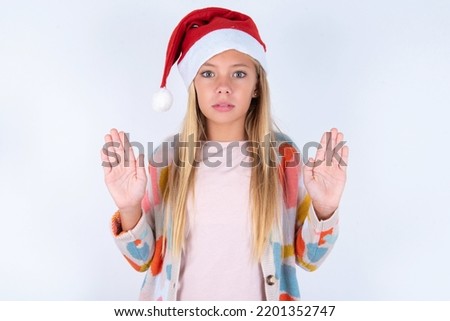 Serious little kid girl with Christmas hat wearing yarn jacket over white background pulls palms towards camera, makes stop gesture, asks to control your emotions and not be nervous