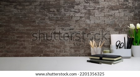 Hipster workplace with flower pot, books and stationery on white table against brick wall.