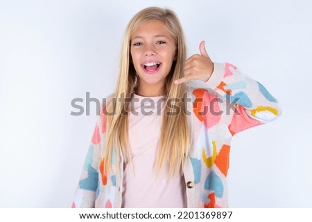 little kid girl wearing colorful yarn jacket over white background makes phone gesture, says call me back again, has glad expression.
