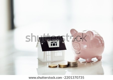 Pink piggy bank with tiny house on white marble background and blurred bokeh with copy space. Concept for financial home loan or money saving for house buying, real estate and insurance