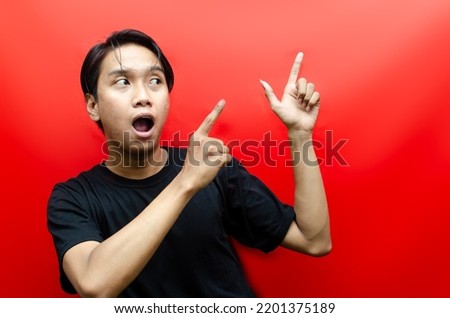 a happy young asian man wears black t-shirt is surprised and shouting wow with pointing his finger to the left isolated on red background. advertising model concept.