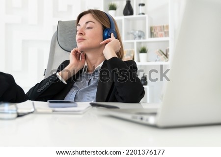 Portrait of successful and attractive business woman in headphones listening to music in her office. Positive female employee in black suit smiling and sitting at the desk in her modern workplace