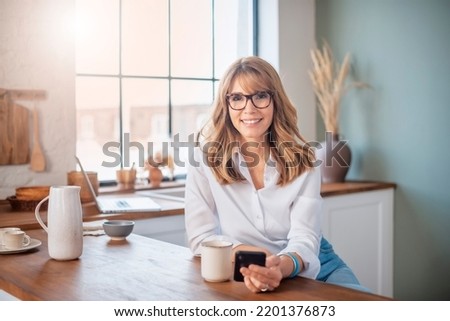 Happy middle aged woman standing in the kitchen at home and text messaging.  Royalty-Free Stock Photo #2201376873