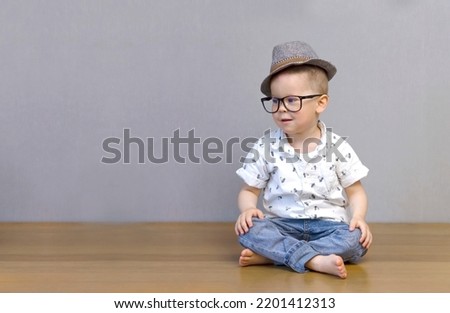 A boy in blue jeans, a blue shirt, a hat and glasses sits cross-legged on the floor, looks away and smiles. High quality photo