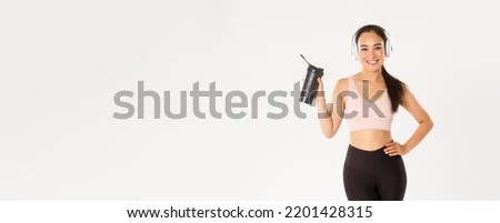 Sport, wellbeing and active lifestyle concept. Attractive slim and fit asian fitness girl in headphones, listening music during workout, drinking water or protein from bottle, white background