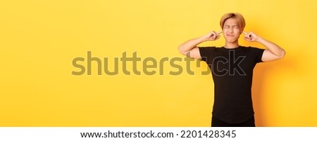 Portrait of annoyed and bothered asian guy grimacing, shut ears disturbed by loud noise, standing yellow background