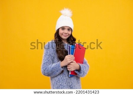 Teenager child in warm sweater and hat hold books and backpack isolated on yellow background. Happy schoolgirl, positive and smiling emotions.