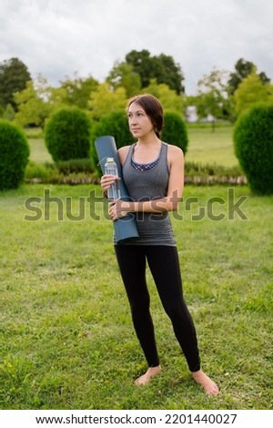Athletic thin young woman holds yoga mat and bottle of water after or before training outdoors in the park cheerful with smile on face with happy and natural expression in sportswear. Vertical photo.