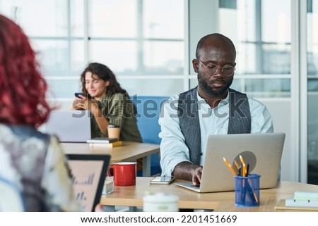 Young serious African American male analyst sitting by desk in front of laptop and organizing work against female coworker using gadgets