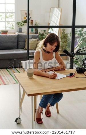 Young brunette woman in white tanktop making notes on paper while sitting by desk at home or radio studio in front of microphone
