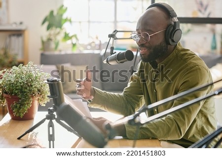 Black guy in headphones talking in microphone while reading plan of discussion and recording new video for online audience