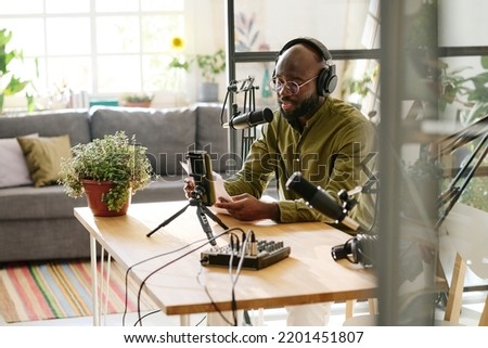 Young male host or blogger reading his notes in microphone while recording new video for online audience in living room